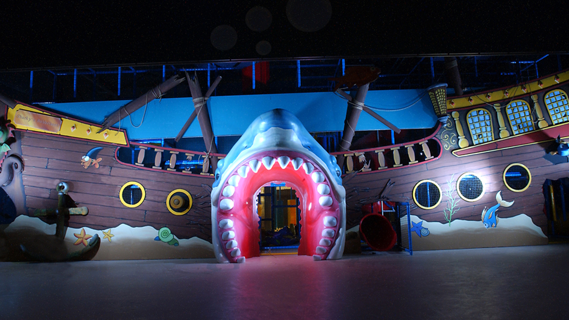 A brightly lit children's play area designed to resemble a pirate ship with a large open-mouthed shark entrance.